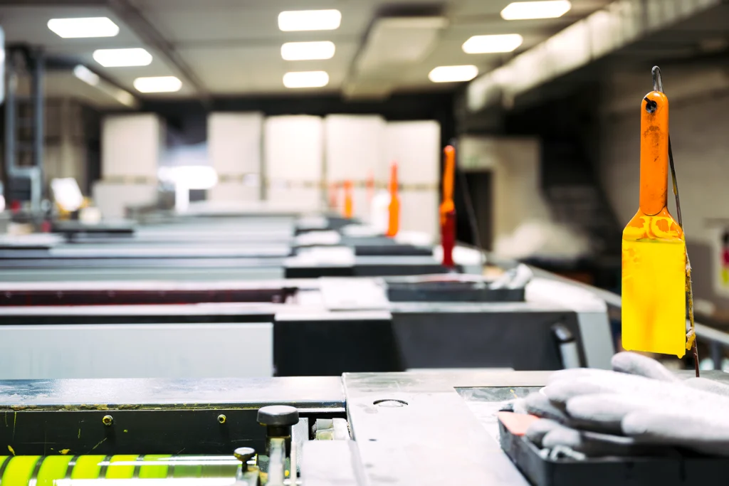 2025_Warlich_Druck_Meckenheim_Januar_JR_1711_300dpi_sRGB Industrielle Druckmaschine in einer Produktionshalle, vorne ein gelbes Farbwerkzeug und eine behandschuhte Hand bei der Bedienung.